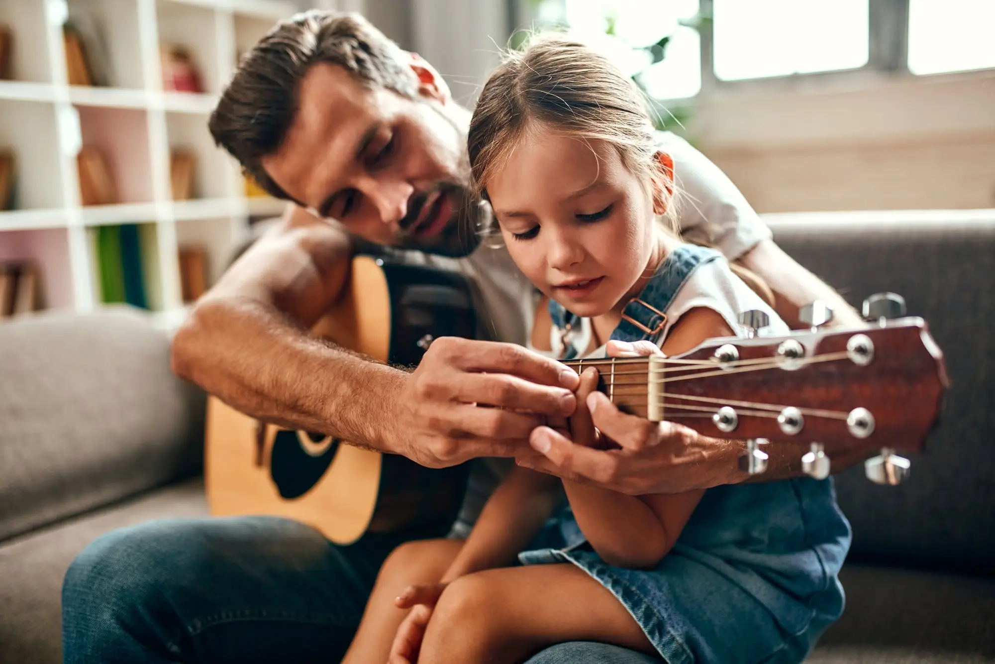 A father on the sofa at home teaching his young daughter to play the guitar